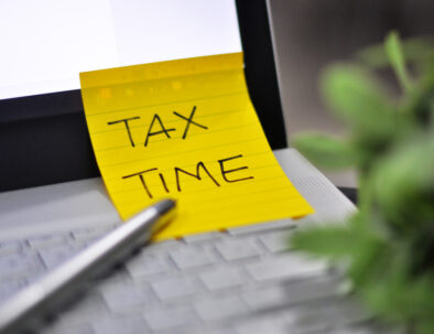 Tax Time: Horizontal top right view of an office laptop background with a metallic pen Tax Time note pad reminder green tree foregrounds - image